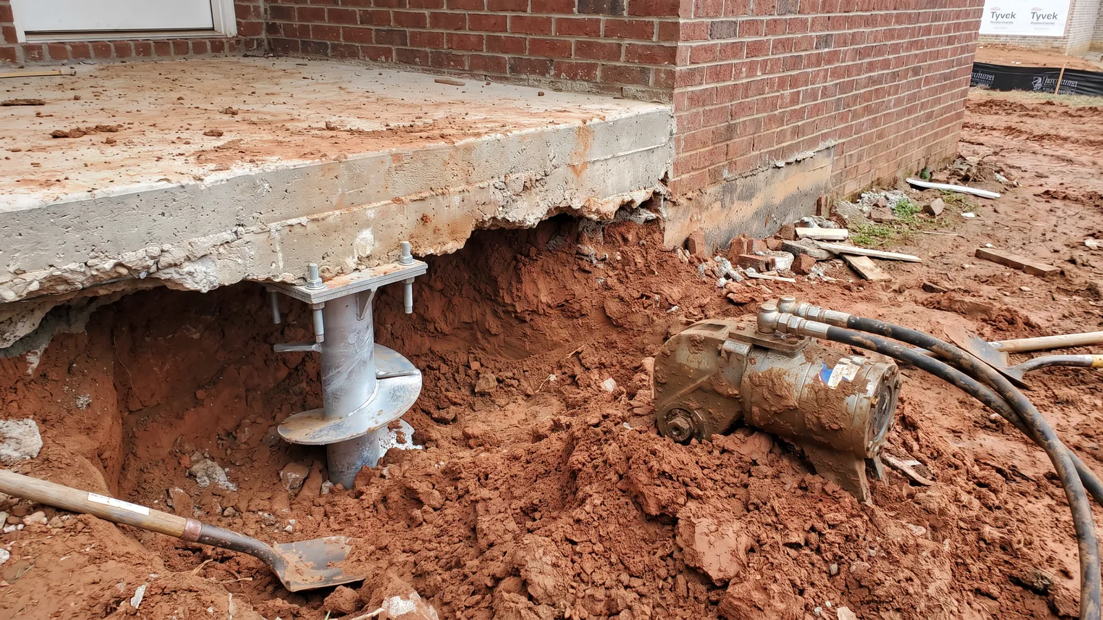 Excavated corner of a concrete slab foundation on a Huntsville-area brick home showing the footing edge, a galvanized helical pier bracket bolted beneath it and a torque motor resting in the hole